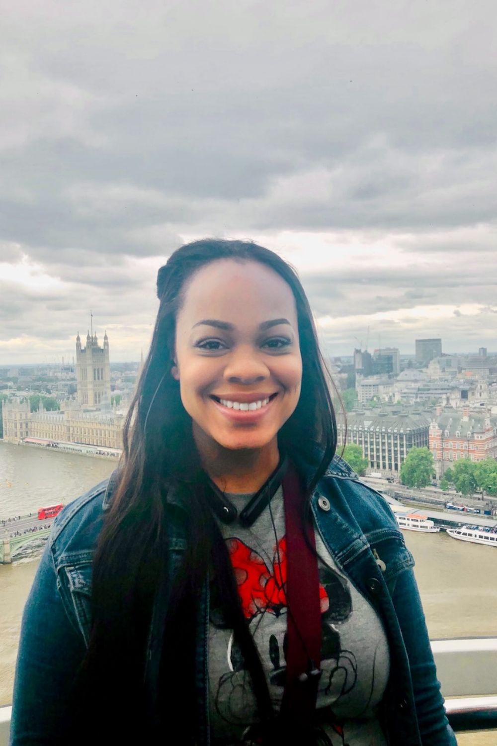 A woman smiles with the iconic Big Ben and the River Thames in the background, enjoying her career break in London, UK. She wears a denim jacket and a Minnie Mouse shirt, exuding happiness and relaxation amidst the city's historic landmarks.