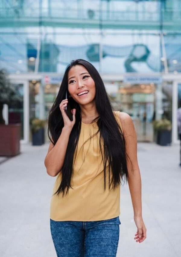 A smiling woman in a yellow top and denim jeans walking while talking on her cellphone, with a modern glass building in the background. Perfect for travelers staying connected with sim cards for France.