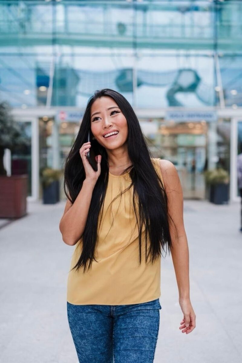 A smiling woman in a yellow top and denim jeans walking while talking on her cellphone, with a modern glass building in the background. Perfect for travelers staying connected with sim cards for France.