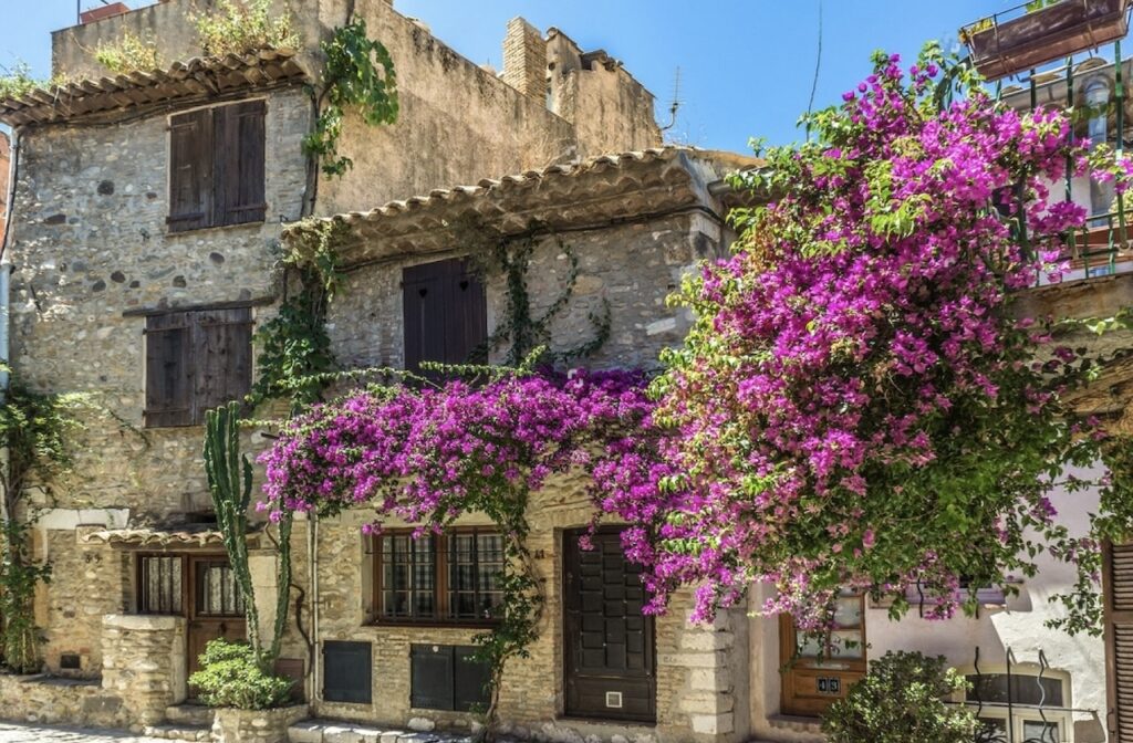 Quaint stone house with rustic wooden shutters, adorned with vibrant bougainvillea flowers in shades of pink and purple, in the charming town of Cagnes-sur-Mer.