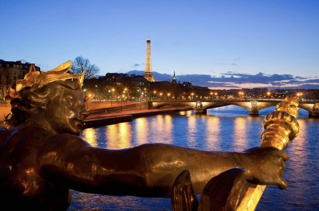 Dusk settles over the Seine River in Paris, framing a bronze sculpture with a gilded laurel wreath in the foreground. In the background, the Eiffel Tower lights up the evening sky, with the Pont des Invalides bridge and its reflections adding to the magical Parisian landscape.