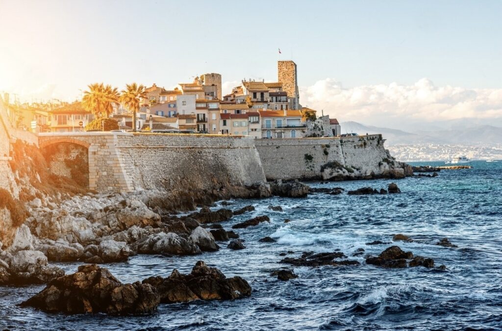 The golden light of sunset illuminates the historic architecture and palm trees of Antibes, set against the backdrop of a rough Mediterranean Sea with rocky shores.