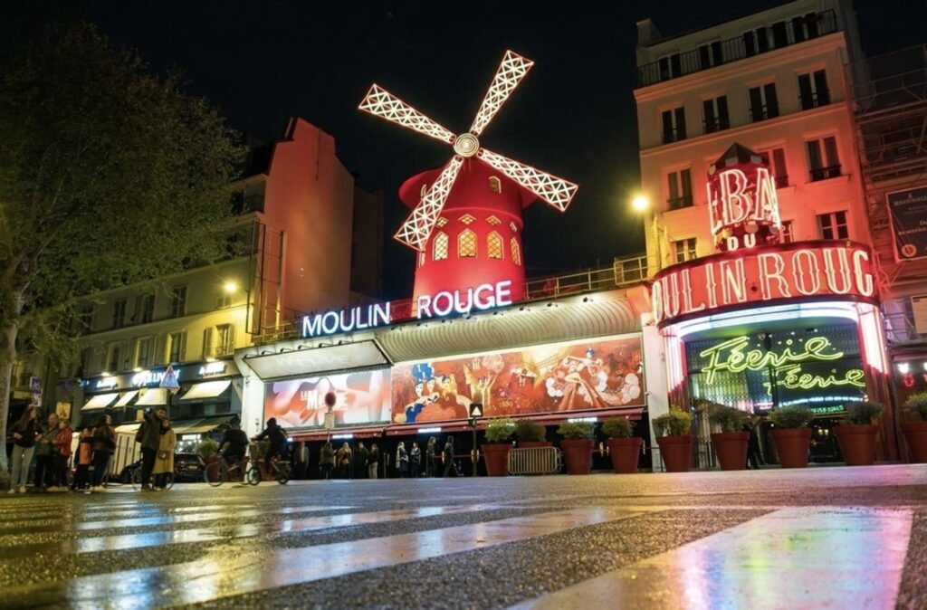 The iconic Moulin Rouge at night, its red windmill lit up against the dark sky, while neon signs add to the lively atmosphere. People gather outside, reflecting the vibrant nightlife of this famous Parisian cabaret venue.
