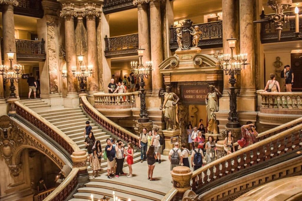 Visitors exploring the grand marble staircase with ornate sculptures and gilded balustrades at the Opéra Garnier in Paris, bustling with activity and architectural grandeur.