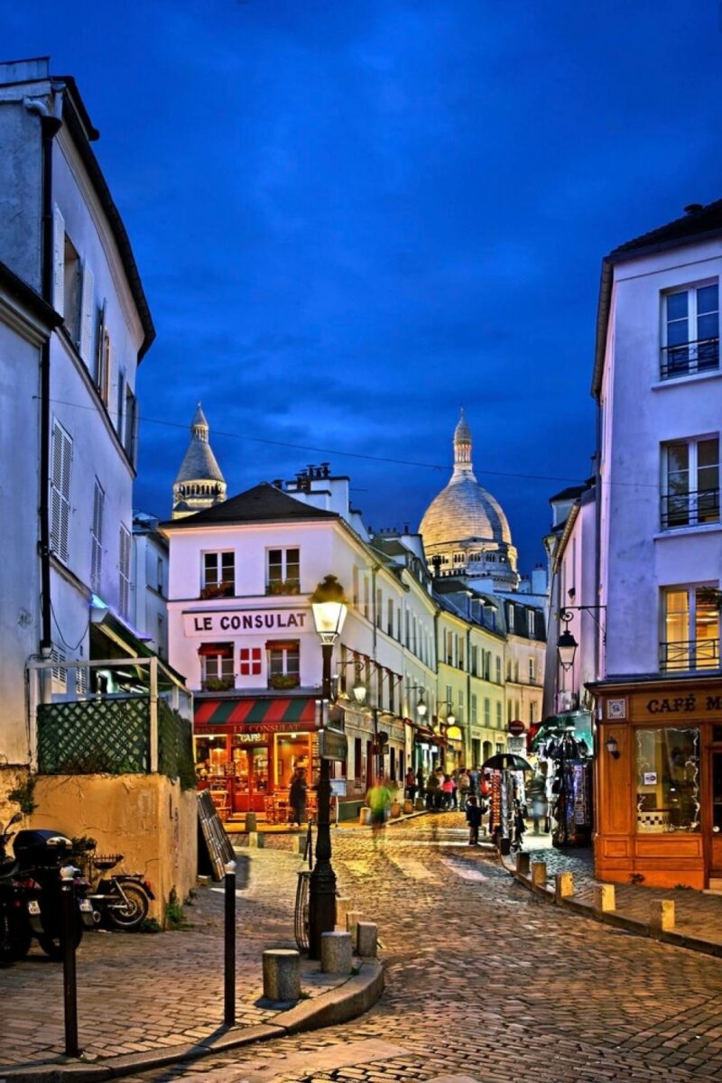Paris at night: a picturesque Parisian street with the illuminated dome of Sacré-Cœur Basilica in the distance. In the foreground, the vibrant red façade of Le Consulat restaurant stands out against the cobblestone street bustling with pedestrians and lined with glowing shopfronts.