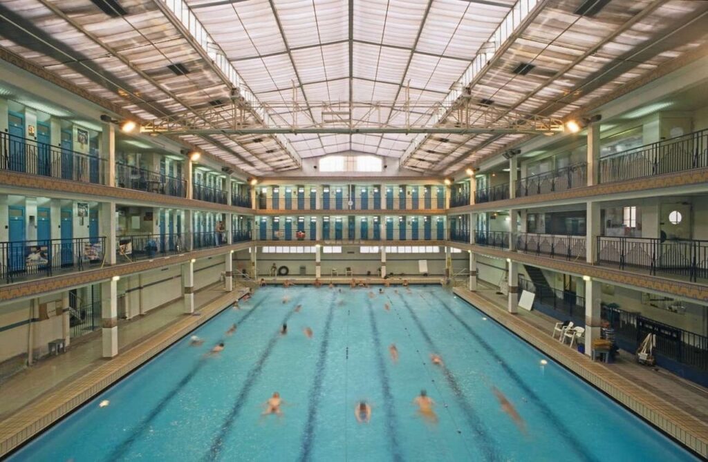 Swimmers glide through the lanes of the historic Piscine Pontoise swimming pool in Paris, characterized by its large, luminous glass roof and double-decker spectator galleries with blue and yellow tiled walls.