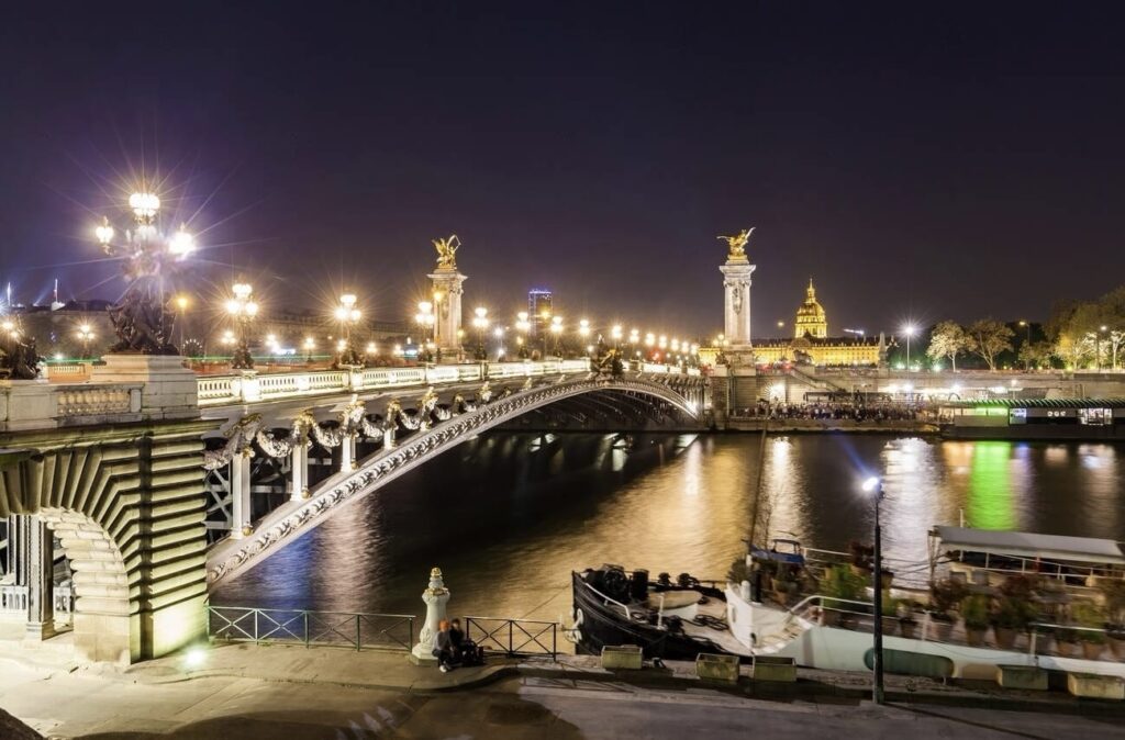 A nocturnal view of the Alexandre III Bridge in Paris, with its ornate lamp posts and statues gleaming under the lights. The Seine River reflects the bridge's lights, and the grand dome of Les Invalides is visible in the background, adding to the city's romantic ambiance.