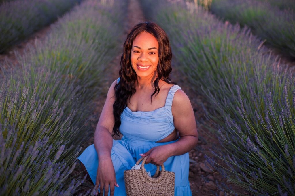 best day trips from Nice: A joyful black woman in a blue sundress sitting amidst a lavender field at dusk in Valensole, France, holding a woven straw bag, with a serene smile on her face.
