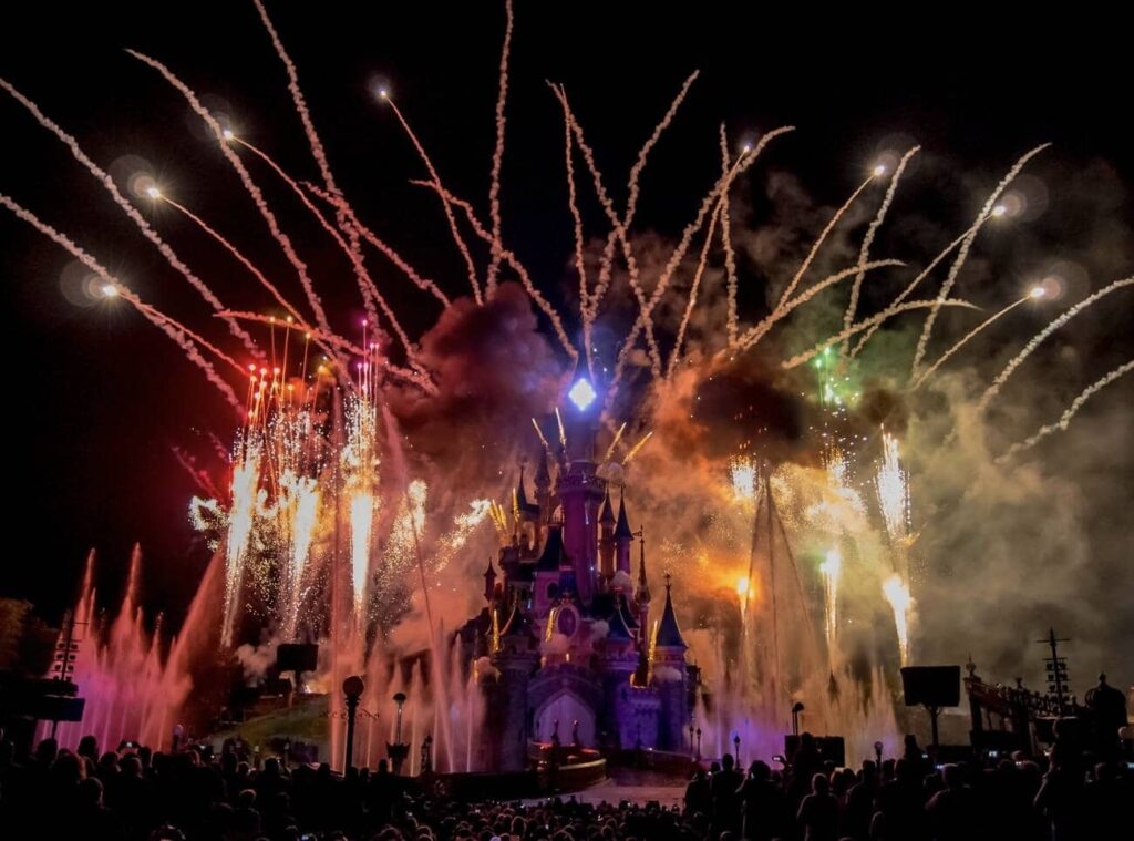 Spectacular fireworks display above a fairy tale castle during a night event, with vibrant bursts of colors lighting up the dark sky. Silhouettes of spectators are visible against the backdrop of the illuminated water fountains and the castle.