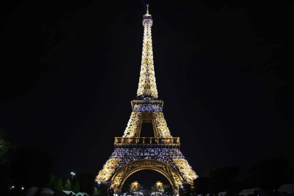 The Eiffel Tower stands majestically against the night sky, bathed in golden light with twinkling lights that make the landmark sparkle. The darkness of the surrounding sky highlights the iconic structure, making it a beacon of Parisian nights.