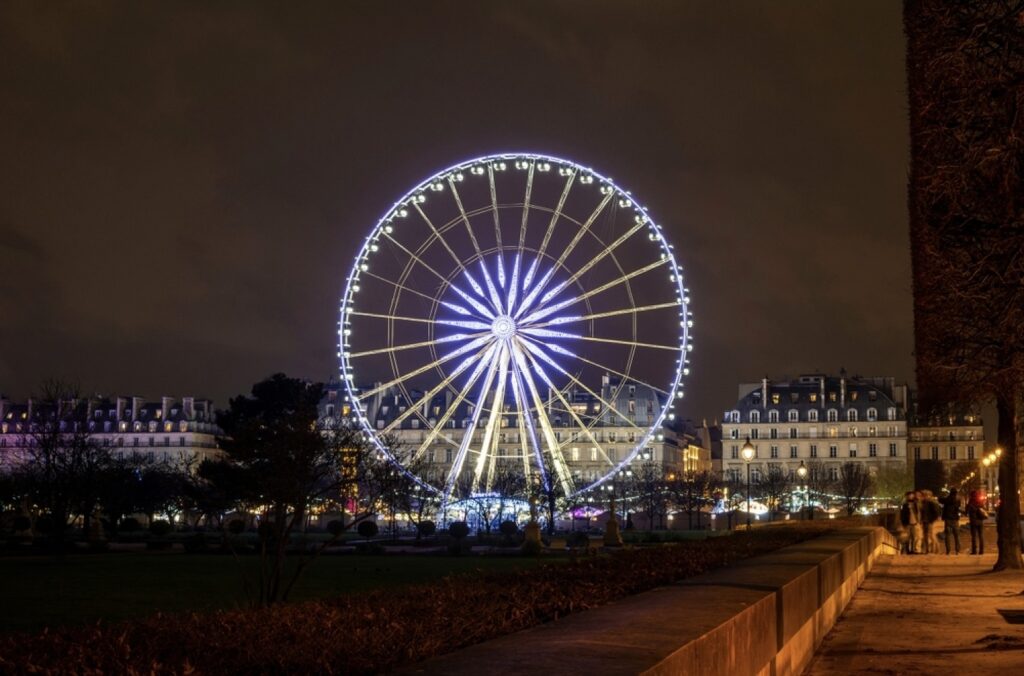 The Ferris wheel in the Tuileries Garden of Paris, France, radiates with bright lights against the evening sky. Onlookers walk along the path, enjoying the view, with Parisian buildings softly lit in the background, creating a picturesque urban scene.