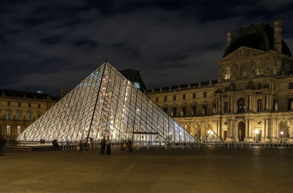 Illuminated Louvre Pyramid at night with the historic Louvre Palace in the background, under a dusky blue sky in Paris.