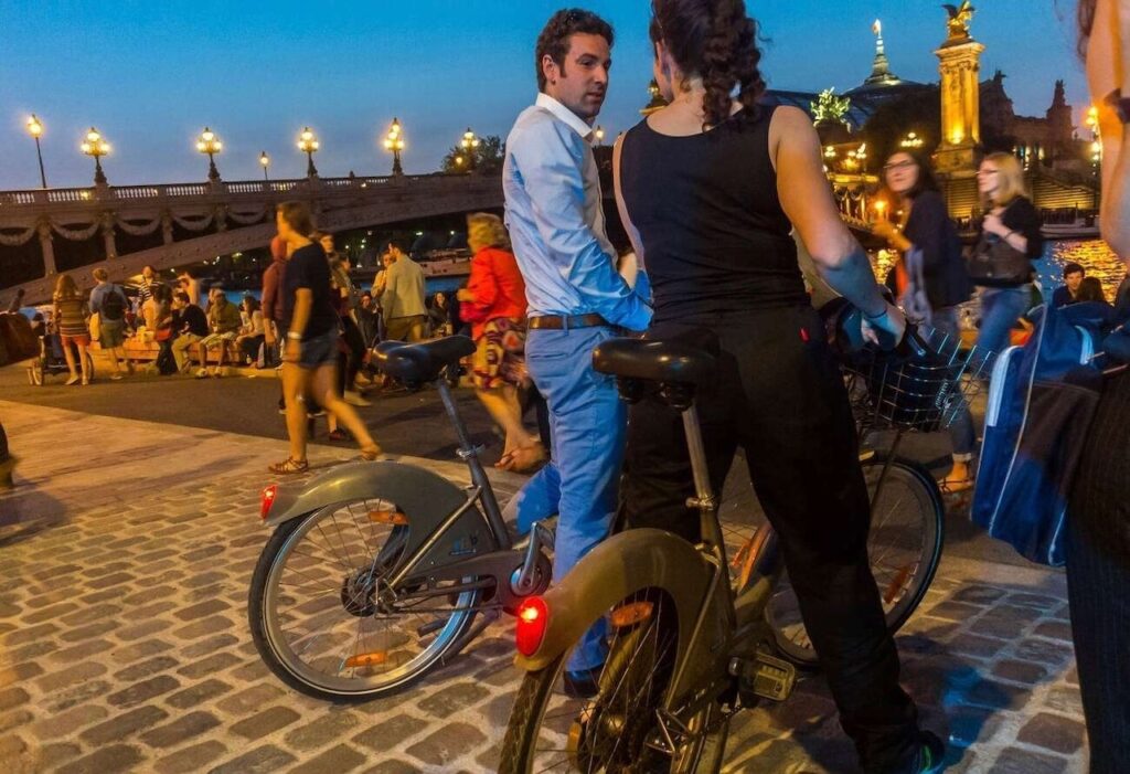 A couple in evening attire pauses on rental bicycles at dusk on a cobblestone path in Paris, with the city's ambient street lights and the lively atmosphere of passersby in the background.