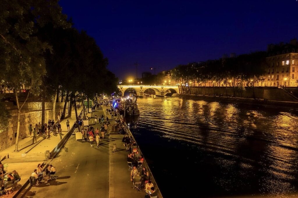 A warm summer night on the banks of the Seine River in Paris, where people enjoy leisurely activities along the promenade. The golden glow of the street lights reflects on the water, with the historical Pont Neuf bridge in the background, adding to the charm of the city's nightlife.