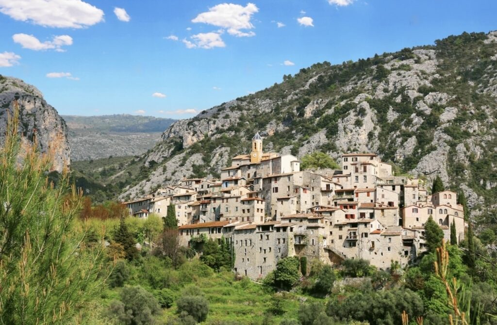The perched village of Peillon, France, with its cluster of traditional stone houses and a bell tower, nestled on the side of a lush green mountain under a blue sky with scattered clouds.
