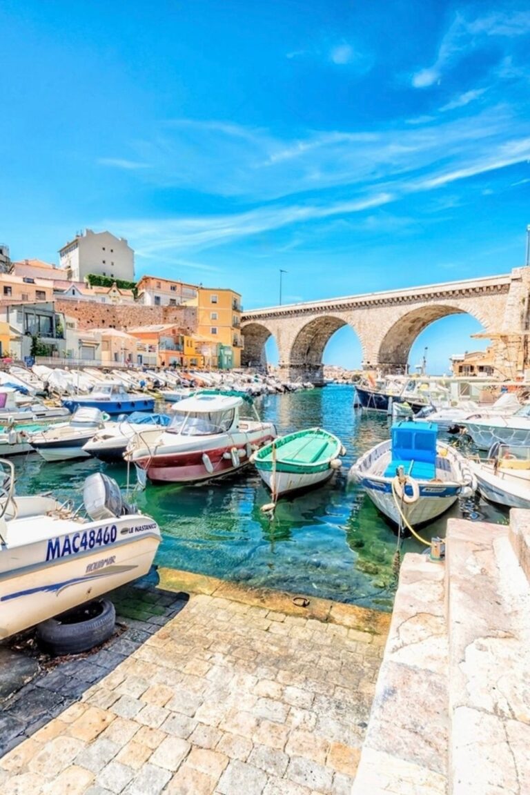 Sunny day at Vallon des Auffes, Marseille, with colorful boats moored in the clear turquoise waters, and the historic stone arch bridge in the background, embodying the charm of Marseille's seaside attractions.