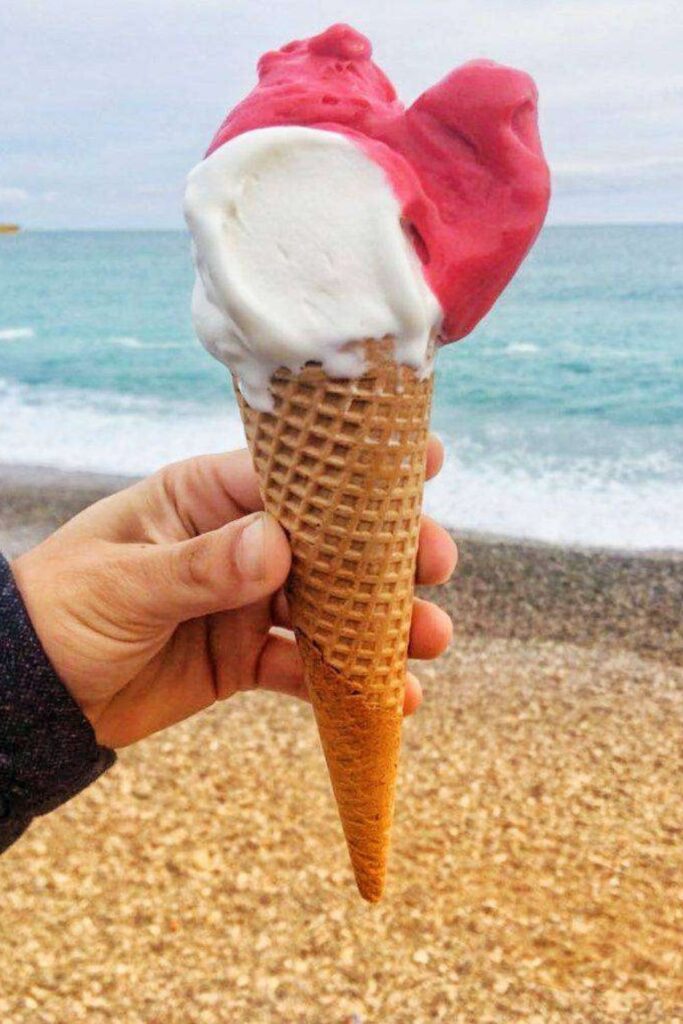 A hand holds up a waffle cone with vibrant pink and creamy white scoops of ice cream against a backdrop of the pebbled beach and turquoise sea, epitomizing a refreshing moment on the French Riviera.
