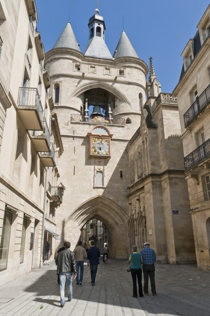 Pedestrians wander towards the iconic Porte Cailhau in Bordeaux, an impressive medieval gate with twin conical towers and a clock, its historical grandeur a stark contrast to the surrounding modern streets, a must-see landmark that is steeped in the city's rich heritage.