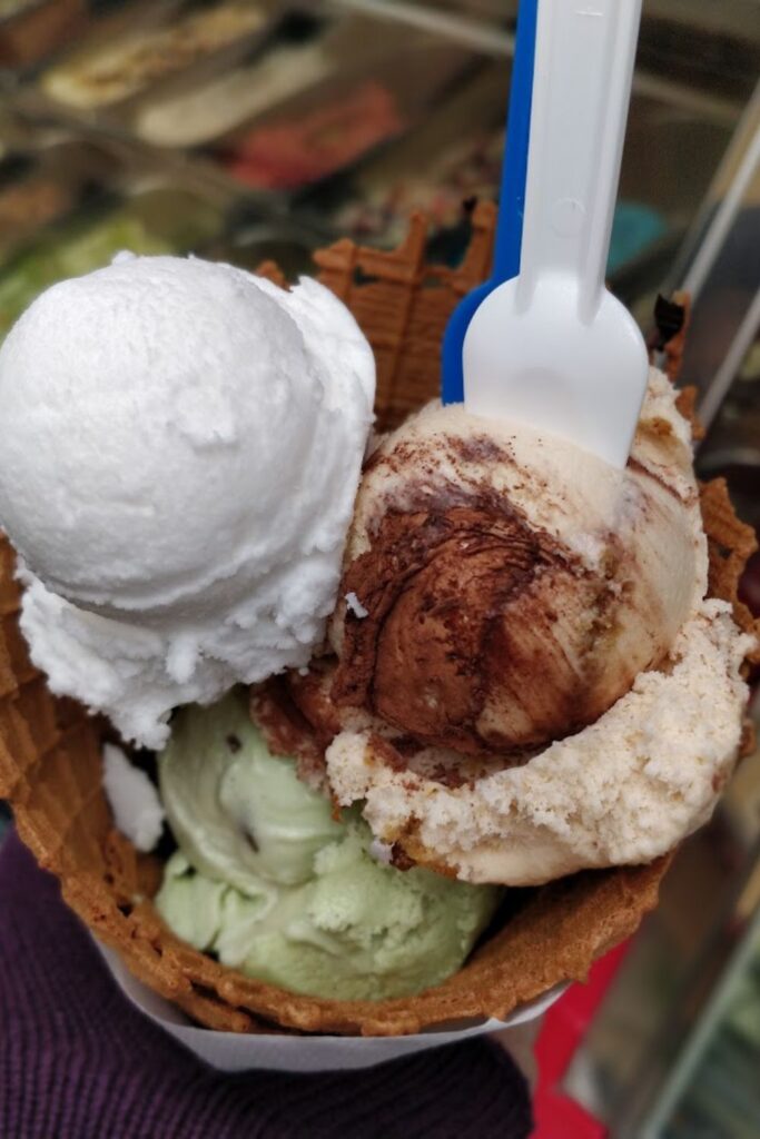 A tempting waffle cone overflows with scoops of ice cream, featuring flavors such as classic vanilla, rich chocolate, and refreshing pistachio, accompanied by a bright blue spoon, encapsulating the indulgent experience at Nice's Roberto 1er.