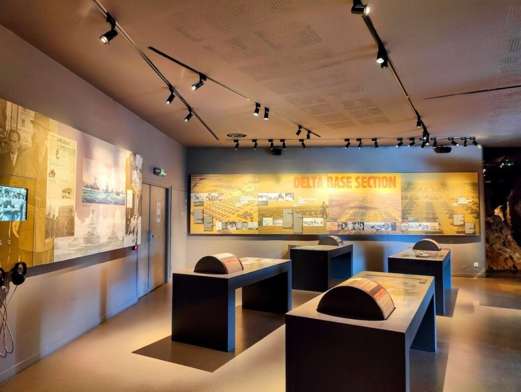 Things to do in Toulon: Interior view of Musée Mémorial du Débarquement exhibit room with the title 'DELTA BASE SECTION' on a wall. The room is lit with track lighting and features large informational panels with text and historical photographs on the walls. In the center, there are several dark rectangular display cases with documents and artifacts. The ambiance is quiet and educational, designed for reflection and learning.