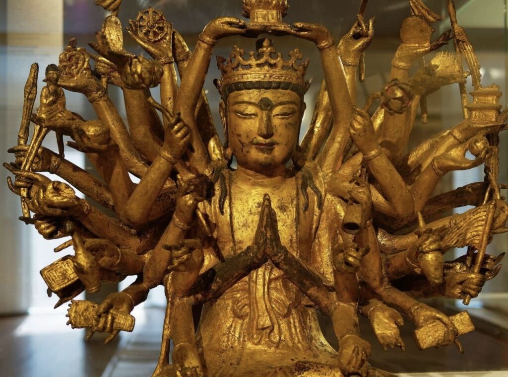 Things to do in Toulon: Close-up of a golden multi-armed statue on display at the Musée des Arts Asiatiques in Toulon, France. The statue depicts a deity with a serene expression, hands in anjali mudra, and multiple arms fanning out, each holding symbolic objects. The intricate detailing and craftsmanship reflect the statue's cultural and religious significance, embodying the artistic heritage of Asian traditions.