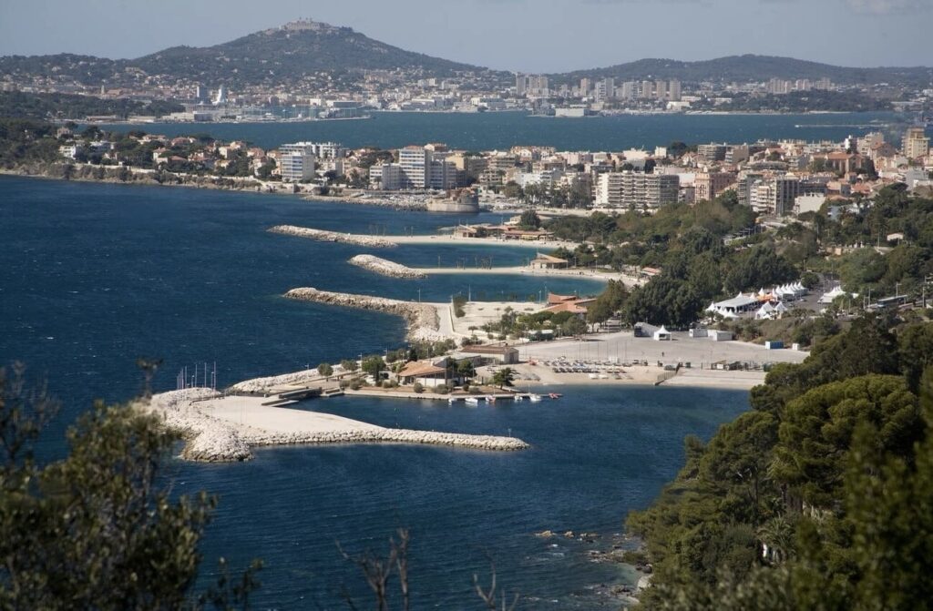 Things to do in Toulon: A sweeping view of the coastline along the Sentier des Douaniers in Toulon, France. The photo captures a clear day with a panoramic view of the Mediterranean Sea, a sandy beach, a marina with several boats, and a series of breakwaters. The surrounding area is dotted with lush greenery, buildings of various sizes, and an urban landscape that rises gently into rolling hills in the background. The atmosphere is serene with natural and man-made elements coexisting harmoniously.