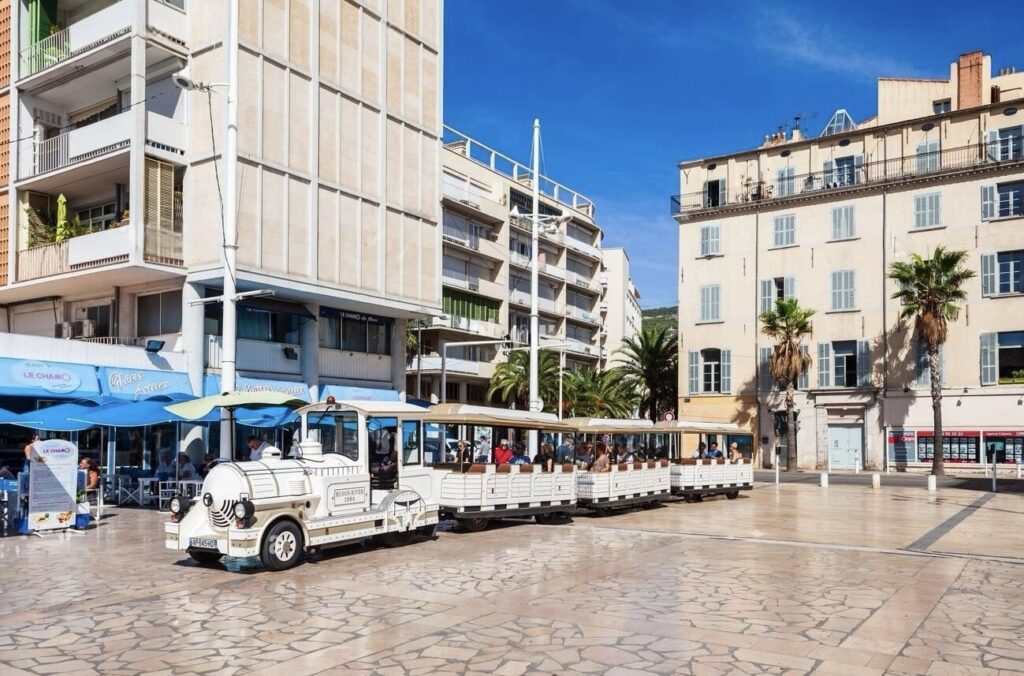 Things to do in Toulon: A tourist train in Toulon, France, with passengers onboard, is parked on a sunny esplanade. The train, designed to resemble a vintage locomotive, is white with blue accents and provides sightseeing tours around the city. It's surrounded by palm trees and urban buildings with restaurants at street level where patrons dine under blue awnings. The atmosphere is lively and leisurely, indicative of a Mediterranean coastal town.