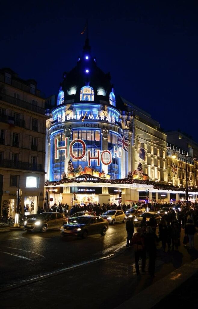 A nighttime view of BHV (Bazar de l'Hôtel de Ville), a famous department store in Paris, France, during the festive season. The building is illuminated with vibrant blue and white lights, accentuating its architectural details and festive decorations, including large 'HO HO HO' lettering draped across the front. The silhouette of the store's dome stands out against the dark evening sky. Crowds of people and busy traffic on the streets reflect the lively urban atmosphere. Holiday shoppers and onlookers gather outside the store, contributing to the bustling Parisian scene.