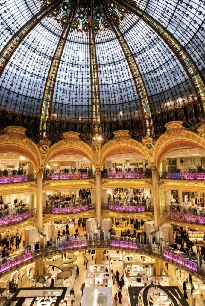 The interior of Galeries Lafayette in Paris, displayed in all its grandeur. A massive, ornate glass dome ceiling casts a warm glow over multiple floors of luxury shopping. The intricate Belle Époque architecture, with its golden, ornamental balustrades and vividly painted pillars, creates a sense of opulence. Shoppers dot the expanse of the department store, browsing through an array of high-end boutiques and designer displays, set against the backdrop of the iconic, lavishly decorated Parisian landmark.