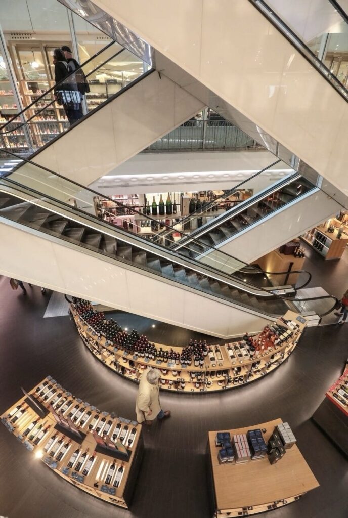 An overhead view inside La Grande Épicerie de Paris, showcasing the sleek design of intersecting white escalators forming an X shape. Shoppers are visible ascending and descending, with one person paused at the convergence point, creating a moment of stillness in the dynamic environment. Below, curved wooden shelves are neatly stocked with an assortment of wines and gourmet foods, inviting exploration. The warm lighting complements the polished floors and enhances the luxurious shopping experience in this renowned Parisian food emporium.