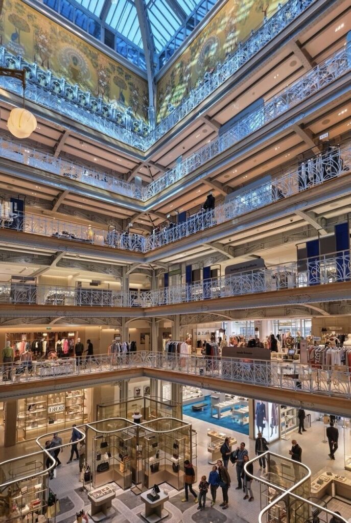 An interior shot of La Samaritaine department store in Paris, showcasing its elegant multi-level architecture. The building features an expansive glass roof that floods the space with natural light, highlighting the decorative frescoes on the upper walls. Ornate wrought-iron railings in white line the numerous balconies that overlook the central atrium. Each floor presents a variety of luxury boutiques and display areas with shoppers casually browsing through. The design combines classic Parisian style with contemporary elegance, creating a sophisticated and inviting shopping experience.