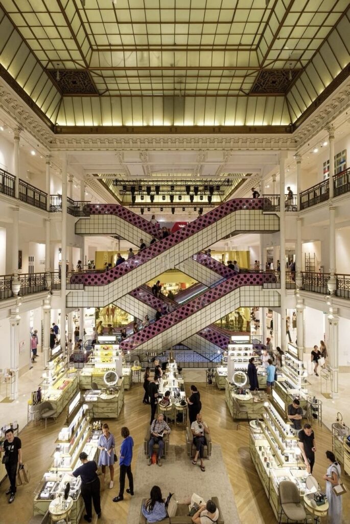 A photograph of the interior of Le Bon Marché Rive Gauche in Paris, featuring the store's distinctive modern escalators with vibrant pink and black polka-dot patterns that crisscross in the center of the image. The classic architecture of the building is highlighted by a decorative white ceiling and ornate metalwork. Below, an array of well-lit cosmetics counters are arranged in neat rows, bustling with shoppers and staff. Customers are seen mingling, shopping, and resting on seats scattered around the space, adding to the lively yet elegant atmosphere of this renowned Parisian department store.