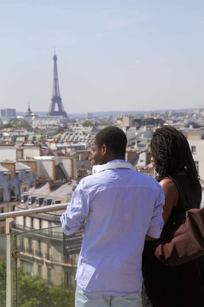 A couple stands on a rooftop terrace with their backs to the camera, overlooking a panoramic view of Paris. The Eiffel Tower looms in the distance under a clear blue sky, symbolizing the romantic and historic essence of the city. The man is dressed in a casual light blue shirt and dark pants, while the woman is wearing a black top and carrying a brown bag, suggesting a relaxed yet stylish ambiance typical of Parisian life.