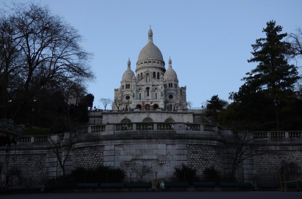 Twilight view of the Sacré-Cœur Basilica, prominently perched atop the Montmartre hill in Paris, with its white domes standing out against the dusky sky, flanked by bare trees and a vintage carousel visible to the left.