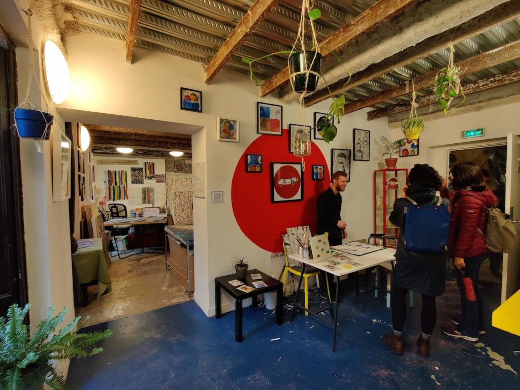 A bustling art studio inside the 59 Rivoli artist collective in Paris, featuring exposed beams and industrial ceiling. The room is filled with an eclectic mix of artwork on the walls and hanging plants, with a prominent red circular motif on the far wall. People are engaged in conversation near a table showcasing prints and various art pieces, creating a lively creative atmosphere.