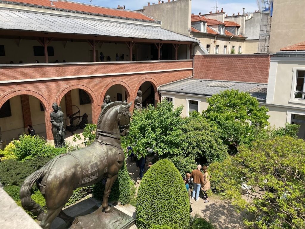 A view overlooking the serene courtyard of the Bourdelle Museum in Paris, featuring a bronze equestrian statue in the foreground. The courtyard is framed by arched brickwork corridors, with visitors strolling among the sculpted greenery and classical statues that contribute to the artistic and tranquil ambiance of the space.