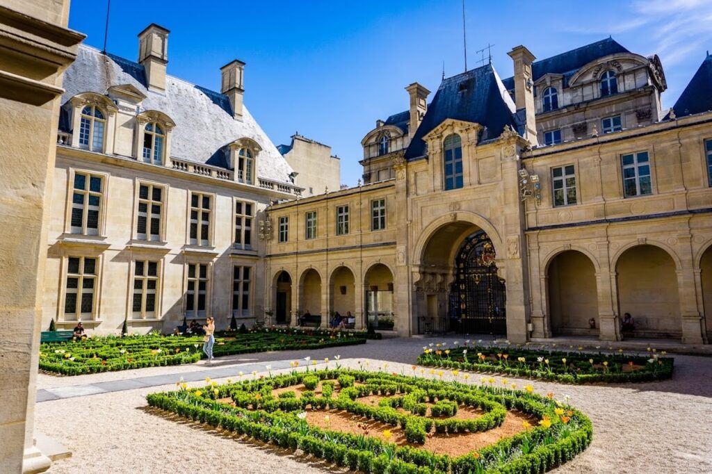 A sunlit courtyard of the Carnavalet Museum in Paris, showcasing manicured garden beds with vibrant flowers in geometric designs. The classical architecture of the surrounding stone buildings, featuring tall windows and elegant archways, frames the peaceful garden setting, where visitors can be seen enjoying the ambiance.