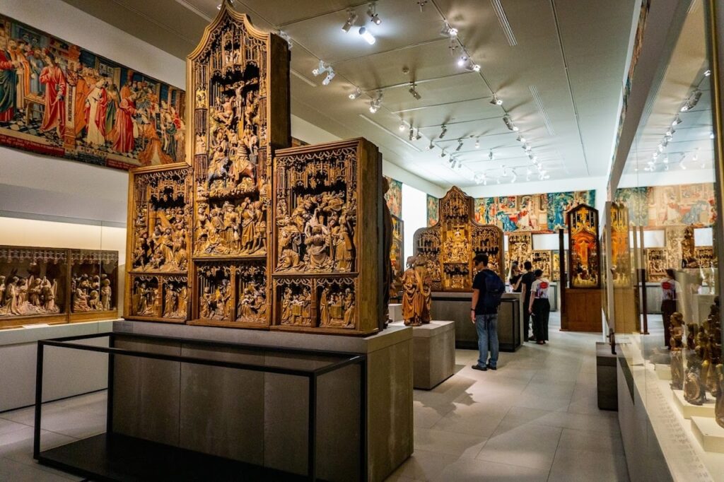Visitors admiring the intricate medieval altarpieces and religious sculptures on display at the Cluny Museum in Paris. The exhibit features detailed wooden carvings depicting biblical scenes, set against a modern backdrop with focused lighting, creating a contrast between the historical artworks and the contemporary museum setting.