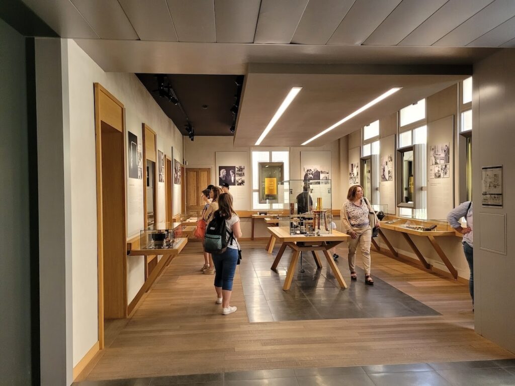 Patrons explore the exhibits at the Curie Museum in Paris, which highlight the scientific achievements of Marie Curie. The gallery is modern and well-lit, with display cases containing historical documents and artifacts, and framed photographs lining the walls, inviting visitors to delve into the legacy of the pioneering scientist.