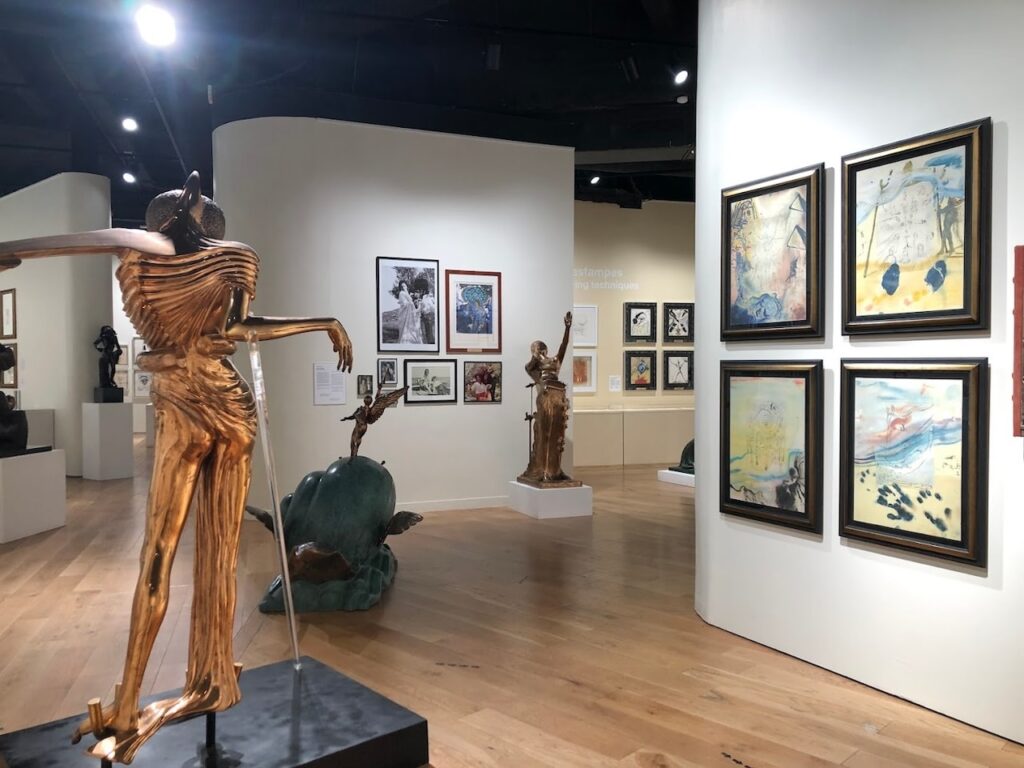 An art exhibit at Dalí Paris, featuring a prominent golden sculpture of a figure with outstretched arms in the foreground. The gallery space is filled with various sculptures and framed artworks, including colorful Dalí paintings on the walls, offering visitors a diverse view of the artist's surreal and imaginative creations.