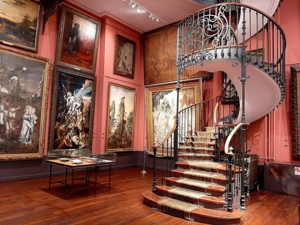 The opulent interior of the Gustave Moreau Museum in Paris, featuring a grand spiral staircase with ornate metal railings. The room is richly decorated with large, framed 19th-century paintings covering the crimson walls, evoking the historic and artistic legacy of the symbolist painter Gustave Moreau.