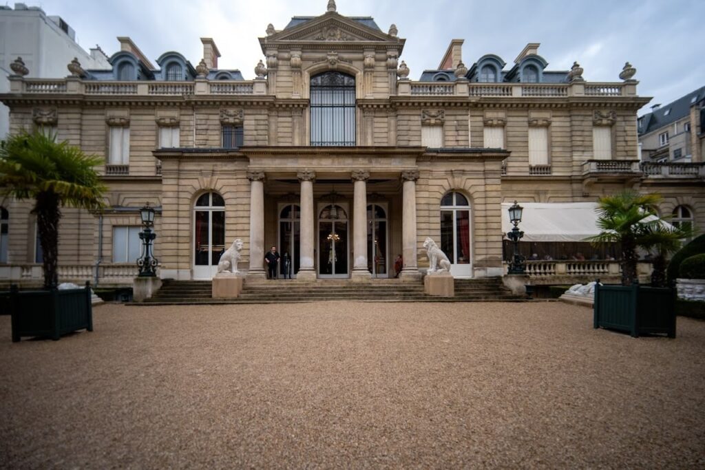 The grand entrance of the Jacquemart-André Museum in Paris, featuring a stately facade with classical architectural details and sculptures of lions flanking the pathway. The elegant building, complete with columns and an ornate lamp post, provides a majestic welcome to visitors stepping onto the gravel forecourt.