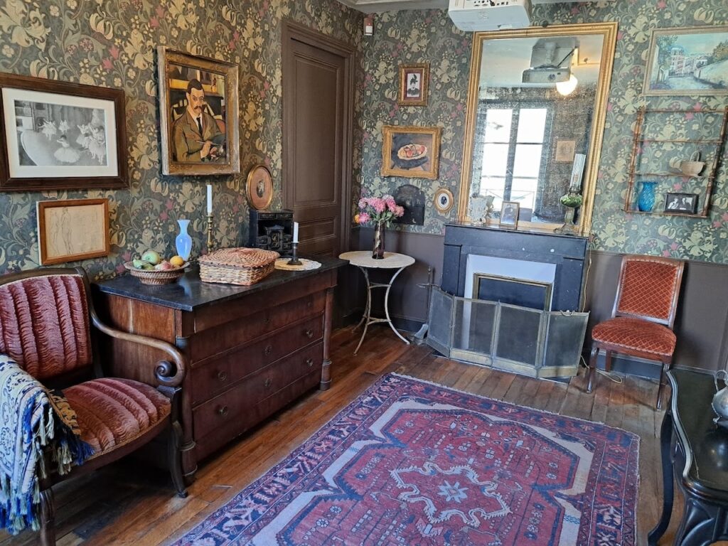 A cozy room in the Montmartre Museum, Paris, decorated with vintage floral wallpaper and period furniture. Portraits and framed pictures adorn the walls, while an ornate mirror above a classic fireplace reflects a warm, inviting atmosphere. The room is complete with a plush red armchair, a well-worn Persian rug, and a side table with fruit, capturing the bohemian spirit of Montmartre.