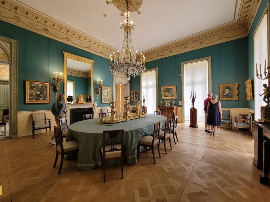 Elegant interior of the Musée Marmottan Monet in Paris, featuring a spacious room with teal walls and classical décor. A crystal chandelier hangs above a large table with a green tablecloth, while paintings in ornate frames, antique furniture, and gilded candelabras add to the room's historical charm. Visitors are quietly observing the art, contributing to the museum's tranquil ambiance.