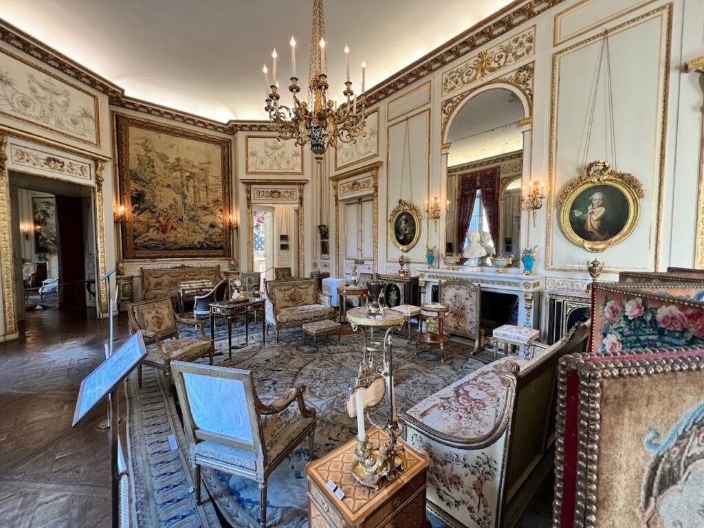 Lavish drawing room in the Musée Nissim de Camondo, Paris, with classical French decor, gilded paneling, and ornate tapestries. The room is furnished with luxurious armchairs, antique tables, and a grand chandelier, complemented by portraits and mirrors, illustrating the opulent lifestyle of the Camondo family in the early 20th century.