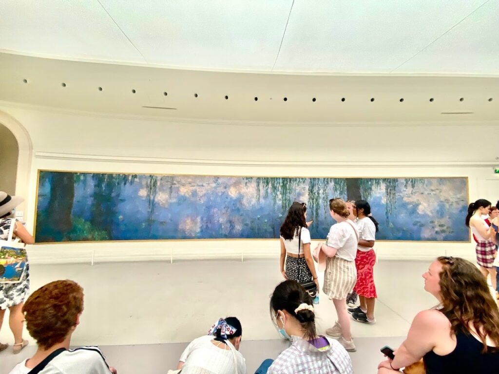 Visitors at the Musée de l'Orangerie in Paris are engrossed in one of Claude Monet's large 'Water Lilies' murals, a panoramic display of his impressionistic water garden in Giverny. The room's curved walls and soft lighting enhance the immersive experience of Monet's tranquil blues and greens.