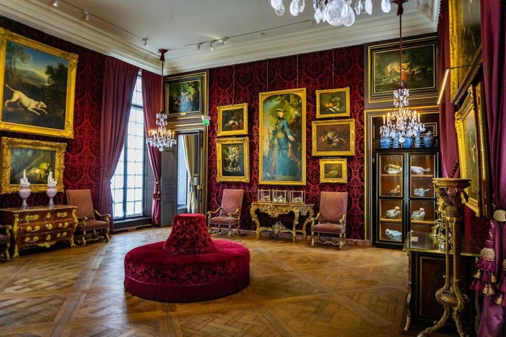 An ornate room within the Musée de la Chasse et de la Nature in Paris, adorned with rich red damask wallpaper and an array of classic paintings in golden frames. The room features a luxurious red velvet settee, a circular centerpiece ottoman, and antique furnishings, with a sparkling chandelier above, reflecting the museum's theme of hunting and nature.