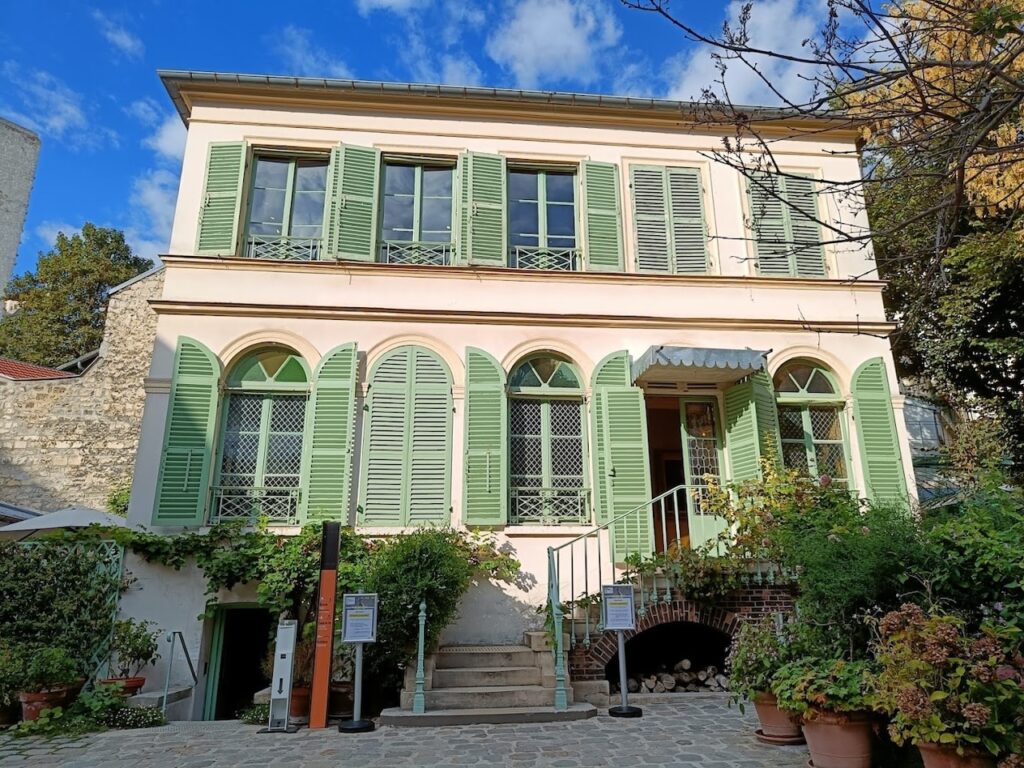 The charming façade of the Musée de la Vie Romantique in Paris, a quaint two-story house with pastel pink walls and contrasting green shutters. The entrance is framed by lush potted plants and a welcoming staircase, evoking the romantic era the museum celebrates.