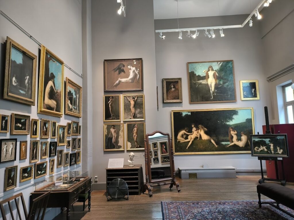 An art gallery within the Musée national Jean-Jacques Henner, Paris, with walls densely hung with paintings of various sizes, predominantly featuring figurative works. The room includes a traditional writing desk, a large mirror, and a Persian rug, creating an intimate setting for viewers to engage with the 19th-century French artist's work.