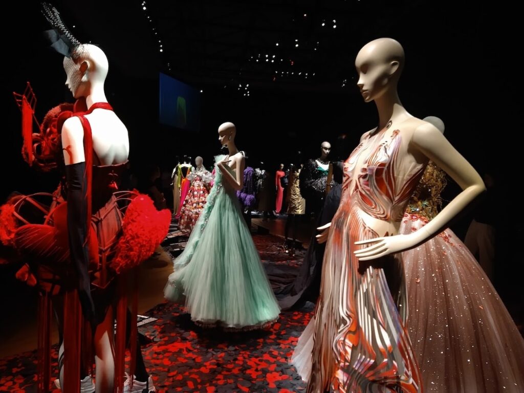 A dramatic fashion exhibition at the Palais Galliera in Paris, with mannequins arrayed in a variety of haute couture dresses from different eras. The dimly lit room, accentuated with spotlights, showcases the intricate designs and fabrics of the garments, creating a captivating visual display against the dark backdrop.